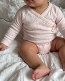 Baby wearing a pale pink long sleeve wrap bodysuit with a small floral print, sitting on a white bed