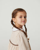 Close up of toddler girl with braided pigtail looking directly at camera, wearing sand striped dress with white collar, studio background
