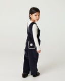 Girl with curly hair wearing navy corduroy overalls and brown cowboy boots, standing in wood paneled room