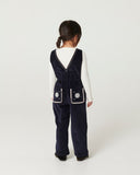 Girl with curly hair wearing navy corduroy overalls and brown cowboy boots, looking up at camera in wood paneled room
