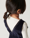 Girl wearing navy corduroy overalls over cream top, standing view from behind showing embroidered sun pockets and dark shoes