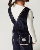 Baby sitting in autumn leaves wearing navy corduroy overalls with embroidered trim over cream sweater, wood fence and grass in background