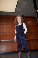 Girl in navy corduroy overalls, side view with embroidered sun pocket visible, standing against plain background