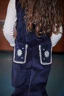 Toddler girl wearing navy corduroy overalls over cream top, close up of embroidered sun pocket detail from behind