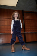 Girl wearing navy corduroy overalls with cream top and dark shoes, standing in wood paneled room, front view