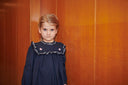 Girl wearing a navy dress with a ruffled bib collar embroidered in bows and pink hearts, standing against a wood paneled wall