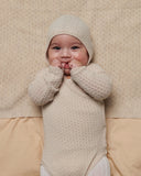 Baby wearing pale cream pointelle knit kimono onesie and matching bonnet, hands clasped at mouth, smiling