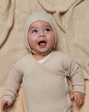 Baby wearing pale cream pointelle knit kimono onesie and matching bonnet, laughing with tongue out, looking upward