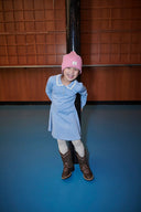 Girl wearing light blue ribbed knit dress with scalloped collar, pink beanie, and embroidered cowboy boots, smiling beside a pole