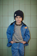 Child wearing a denim jacket, gray shirt, and black beanie against a green tiled wall.