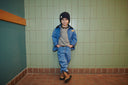Boy wearing denim jacket with navy corduroy collar over grey sweater and jeans, leaning against green tiled wall, brown boots