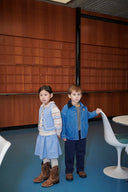 Girl in light blue quilted vest and knit dress holding hands with boy in denim jacket, wood paneled room with round table