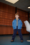 Boy wearing denim jacket with corduroy collar over mustard sweater and navy pants, standing in wood paneled room