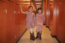 Two girls holding hands in wood paneled hallway, one in pink corduroy overalls with brown cowboy boots, one in pink striped dress with white tights