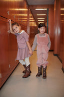Girl in pink striped dress leaning against locker holding hands with girl in pink corduroy overalls, wood paneled hallway