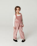 Girl in pink corduroy overalls with embroidered bow pocket and black mary jane shoes, standing in studio