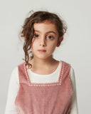 Girl leaning against wood paneled wall wearing pink corduroy overalls over brown knit top, side view showing embroidered bow pocket