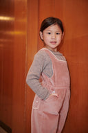 Girl in pink corduroy overalls and brown knit top standing in wood paneled hallway, hand in pocket