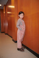 Girl in pink corduroy overalls and brown knit top leaning against wood paneled wall, looking over shoulder at camera