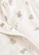Close-up of a baby garment in warm off-white fabric with a scattered teddy bear print in beige and tiny light speckles, plus visible snap buttons.