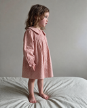 A young girl with wavy brown hair stands barefoot on a bed wearing a dusty pink long-sleeve nightgown with a ruffled collar and subtle polka dot pattern, set against a plain gray wall.