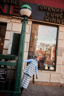 Girl leaning against a lamppost outside a New York diner, wearing cream apron dress with blue floral stripe print over a light blue turtleneck