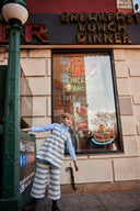 Toddler in cream and blue floral stripe pants and apron dress, leaning against a lamppost outside a diner with BBQ signage