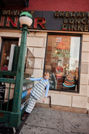 Girl leaning against a green lamppost outside a diner, wearing cream apron dress with blue floral stripe print over a light blue turtleneck