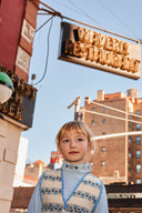Girl standing under a restaurant sign, wearing cream apron dress with blue floral stripe print over a light blue turtleneck, city buildings in background