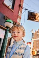 Close up portrait of girl wearing cream apron dress with blue floral stripe print and light blue turtleneck, diner signage in background