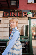 Girl in cream apron dress with blue floral stripe print, walking past a diner storefront, worn over a light blue turtleneck