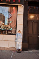 Girl standing against a dark doorway, wearing cream apron dress with blue floral stripe print over a light blue turtleneck, evening light