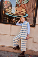 Toddler in cream and blue floral stripe pants and apron dress, resting head on hand against a city storefront window