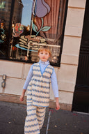 Toddler in cream and blue floral stripe pants and matching apron dress, standing in front of a diner window with a pancake mural