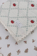 Close up of folded baby blanket corner with sage and cream checked weave, embroidered red apples and scalloped lace trim, Bebe Organic label showing