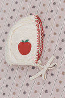 Close up of cream sherpa apple bonnet with red apple embroidery and red blanket stitch trim, flat lay on snowflake print fabric