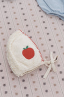 Cream sherpa apple bonnet next to pale blue romper, flat lay on snowflake print fabric