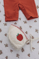 Cream sherpa apple bonnet paired with rust colored pants, flat lay on floral fabric