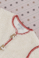 Close up of cream sherpa coat neckline with red blanket stitch trim and wooden toggle closure, Bebe Organic label visible, on snowflake print backdrop