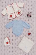 Cream sherpa coat with red apple applique pockets, matching apple bonnet, light blue corduroy romper, patchwork blanket, and felt doll and heart, flat lay on snowflake print