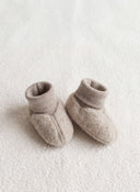 A pair of soft knit baby booties in a light taupe-beige color, displayed on a textured cream fleece surface.