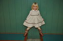 A young girl with blonde hair stands in a dressy outfit consisting of a cream-colored textured dress with dark brown trim, a white Peter Pan collar, and brown cowboy boots, posing against a green wood-paneled wall.
