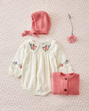 A baby clothing set laid flat on a light pink floral background, including an off-white long-sleeve blouse with floral embroidery, a pink knit bonnet, a pink knit cardigan, and a pink flower hair clip.