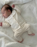Sleeping newborn on white linen bedding wearing pearl cashmere cotton cardigan and pants set with embroidered floral collar, top down view