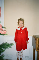 Toddler girl wearing red knit dress with cream scalloped collar, standing by Christmas tree in patterned tights