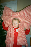 Toddler girl wearing red knit dress with cream collar, oversized red gingham bow on head, standing by Christmas tree