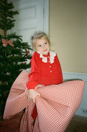 Toddler girl wearing red knit dress with cream collar, holding large red gingham bow, standing by Christmas tree