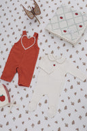 Rust red overall styled flat lay with a cream apple patchwork blanket, cream footed romper with apple embroidery, and a wooden toy horse