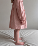 A child wearing a dusty pink long-sleeve dress with a small repeating floral pattern, standing barefoot on a bed with striped bedding against a plain wall.