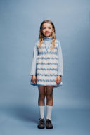 Full length studio portrait of girl in cream apron dress with blue floral stripe print, light blue turtleneck, white socks and brown shoes, looking up and to the side